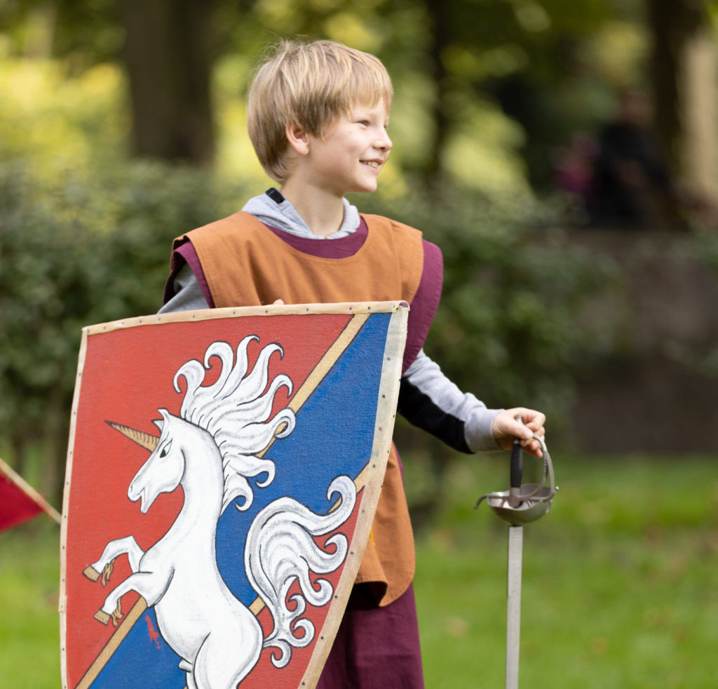 Junge auf einer Wiese in mitteralterlicher Robe hält ein Ritterschild und ein Schwert