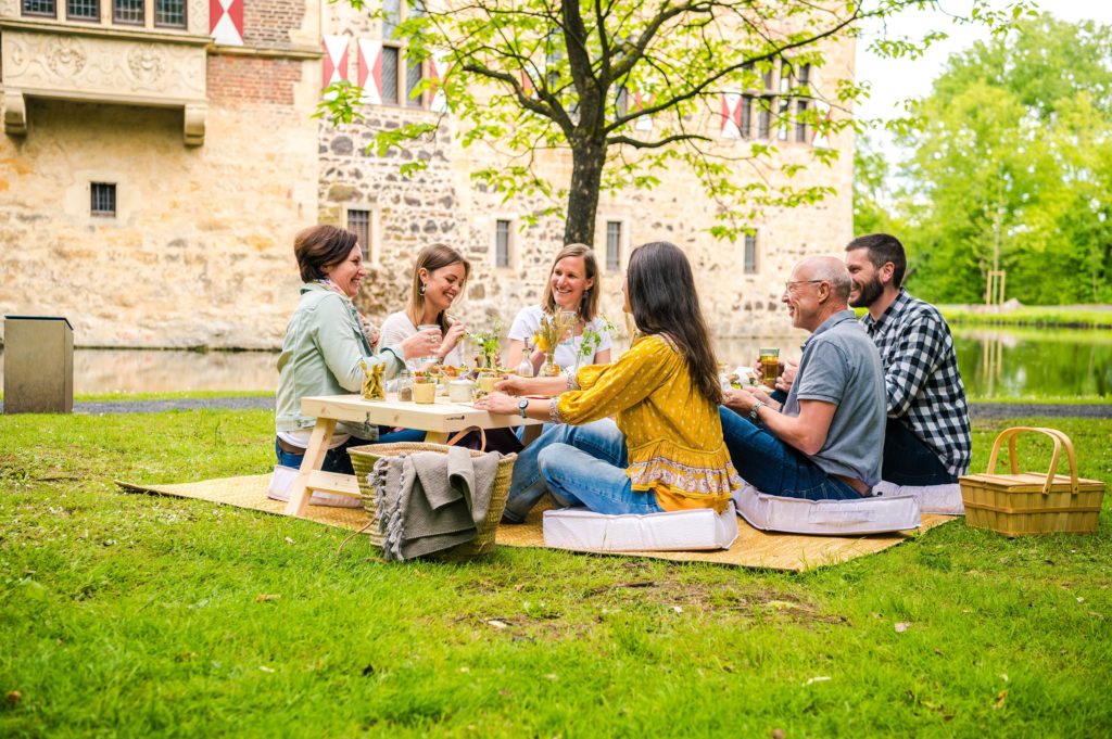 Eine Gruppe von sechs Menschen sitzt auf einer Picknickdecke auf der Wiese vor der Burg Vischering.