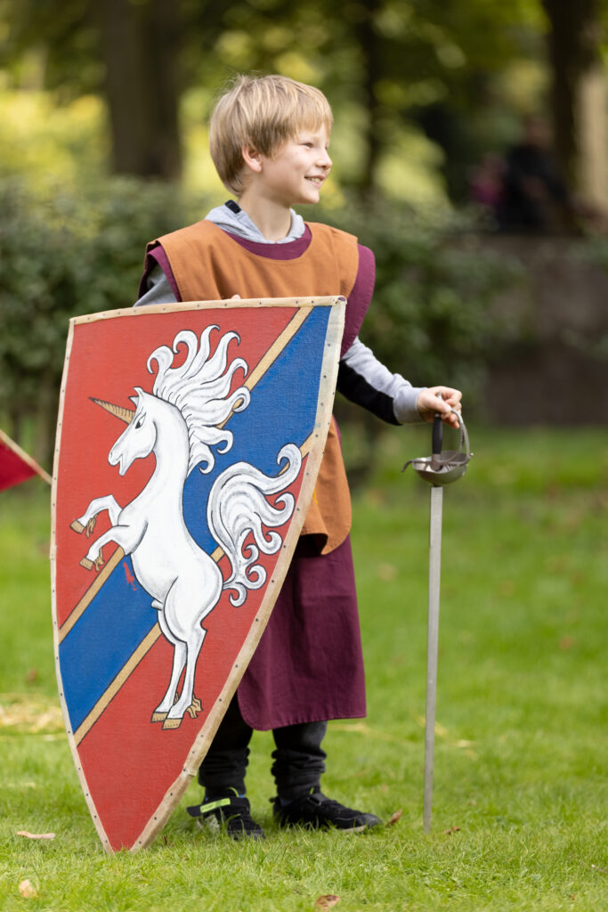 Ein Junge ist als Knappe verkleidet und hält ein Schild mit einem Einhorn sowie ein Schwert.