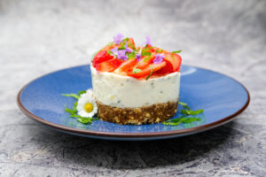Auf einem blauen Teller steht ein rundes Dessert mit Nussboden, cremiger weißer Schicht und frischen Erdbeeren obenauf. Es ist mit Minze, violetten Blüten und einem Gänseblümchen dekoriert.