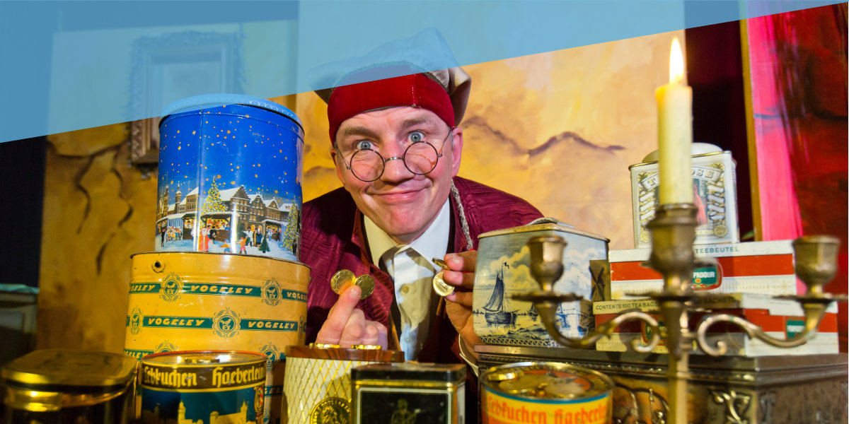 Ein Mann mit Brille und roter Mütze steht vor einem Hausfen Keksdosen und hält Goldmünzen in der Hand.